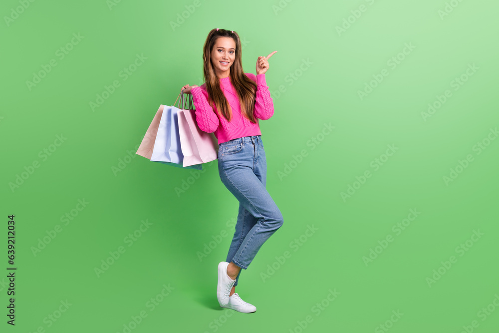 Full length photo of optimistic woman dressed pink sweater indicating at sale empty space hold clothes isolated on green color background