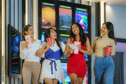 A group of multiethnic friends walk in front of the cinema holding glasses and popcorn having fun. photo