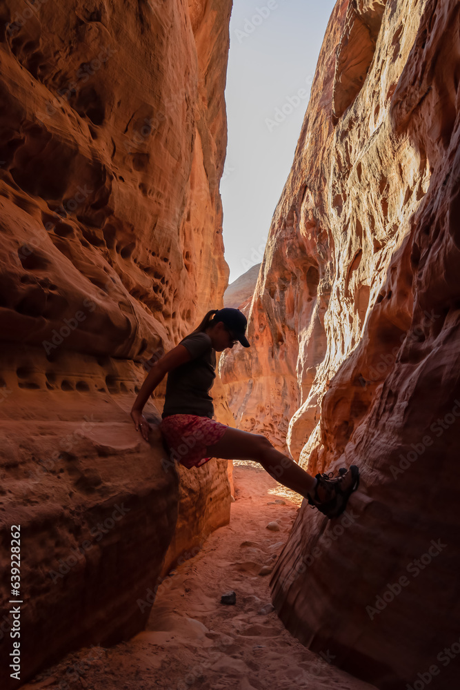 Woman climbing the walls in narrow Kaolin Wash slot canyon on White Domes Hiking Trail in Valley of Fire State Park in Mojave desert, Nevada, USA. Massive cliffs of striated red white rock formations