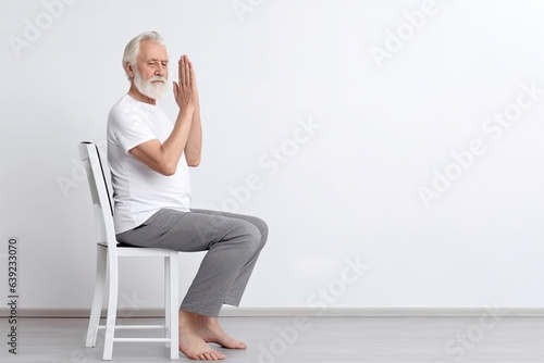An elderly man practices yoga asana or sports exercises for legs and arms on a chair. The concept of sports in old age, exercise therapy, rehabilitation. Banner. Copy space