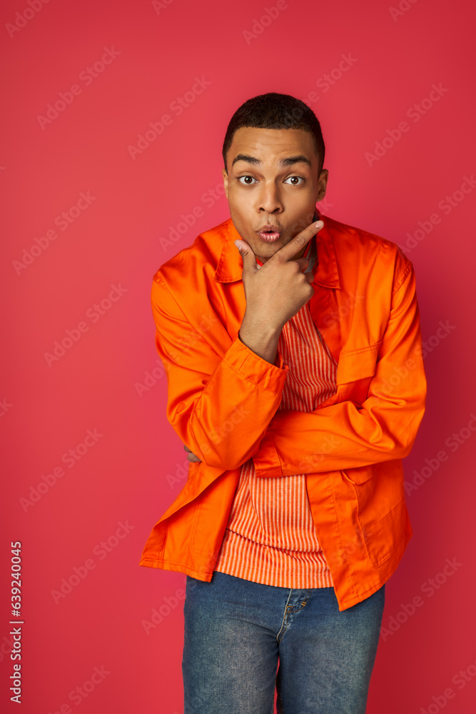 thrilled african american man in orange shirt, with hand near face looking at camera on red thrilled african american man in orange shirt, with hand near face looking at camera on red