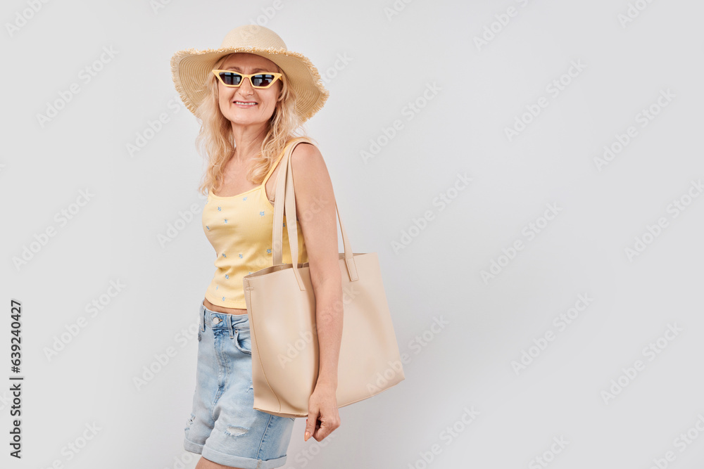Portrait of happy beautiful blonde mature woman in yellow summer clothes, straw hat, sunglasses and shopping bag smiling isolated on white studio background. Vacation and travel concept Portrait of happy beautiful blonde mature woman in yellow summer clothes, straw hat, sunglasses and shopping bag smiling isolated on white studio background. Vacation and travel concept