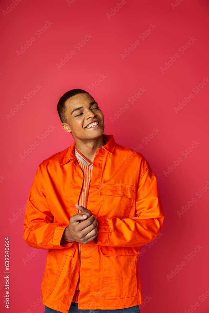 joyful and trendy african american man in orange shirt looking away on red background