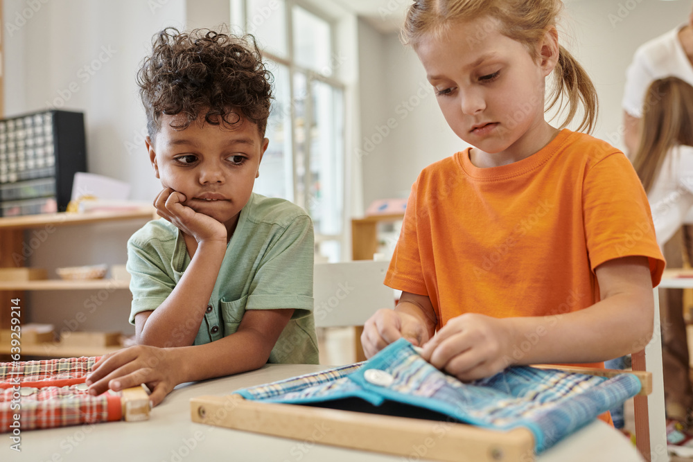 african american boy looking at friend playing with cloth and buttons in montessori school african american boy looking at friend playing with cloth and buttons in montessori school