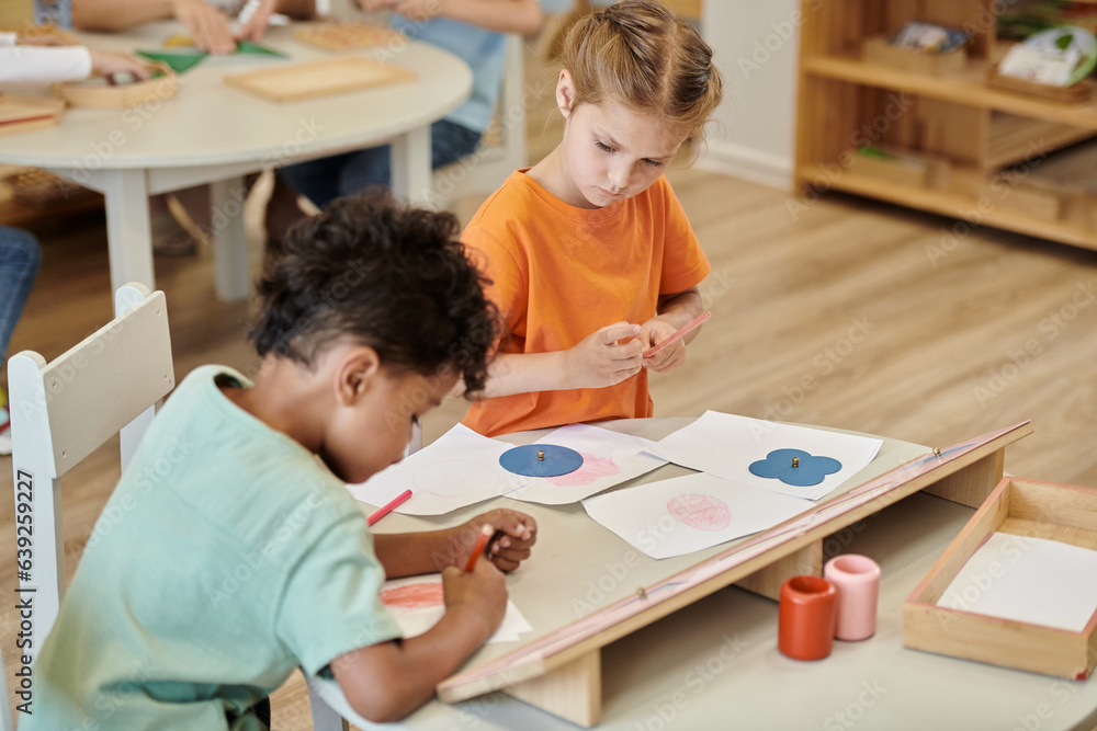 multiethnic children drawing with pencils on table in class of montessori school multiethnic children drawing with pencils on table in class of montessori school