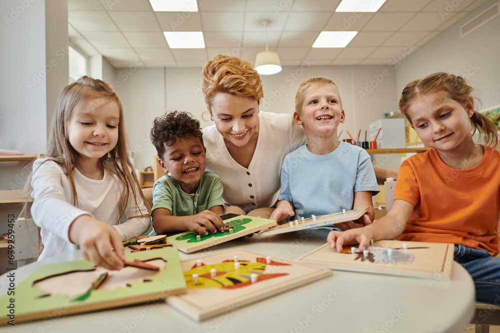 cheerful teacher sitting near multiethnic kids playing with didactic material in montessori class cheerful teacher sitting near multiethnic kids playing with didactic material in montessori class