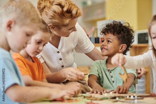 teacher talking to smiling african american boy while kids playing in montessori school