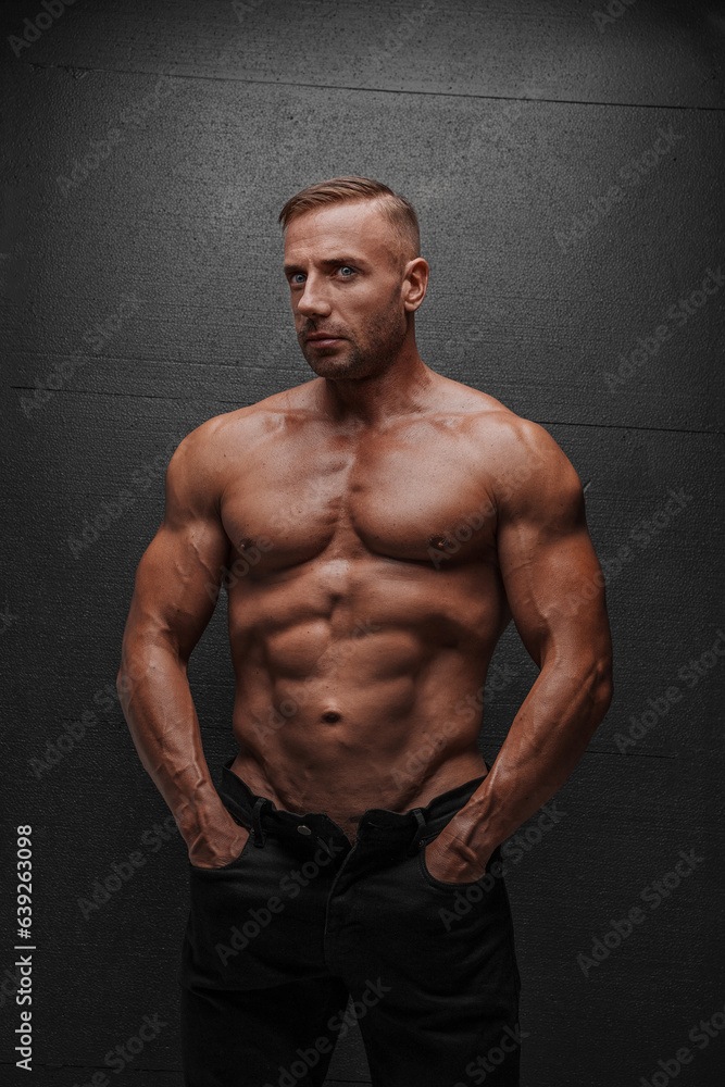 Fototapeta premium Intense and captivating image of a handsome man with a sculpted body, in a provocative pose, wearing black jeans against a black backdrop