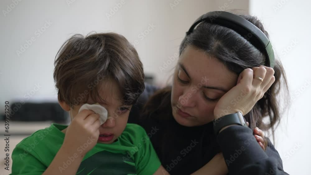Tearful child holding cotton into hurt eye with patient but tired mother waiting for emergency service at hospital Tearful child holding cotton into hurt eye with patient but tired mother waiting for emergency service at hospital