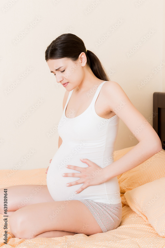 Young pretty pregnant woman, in pain, sitting on bed in bedroom, preparing to give a birth Young pretty pregnant woman, in pain, sitting on bed in bedroom, preparing to give a birth
