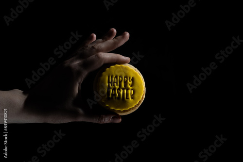A hand holds a cookie with Happy Easter written on yellow icing against a black background, studio lighting