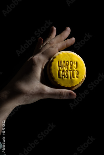 A hand holds a cookie with Happy Easter written on yellow icing against a black background, studio lighting