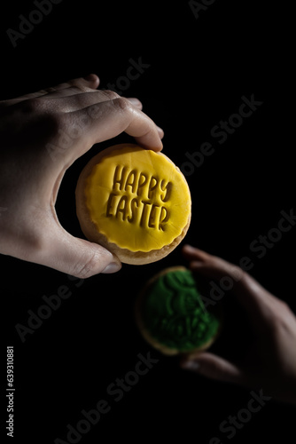 Two individuals hold two cookies with green and yellow icing, reading Easter in the foreground, with a blurred rabbit design
