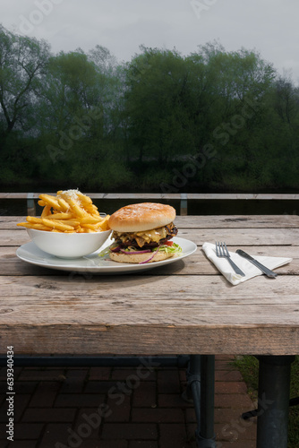 A tasty burger with a juicy meat patty and veggies, accompanied by a portion of French fries, placed on a table at a riverside café, with fork and knife nearby