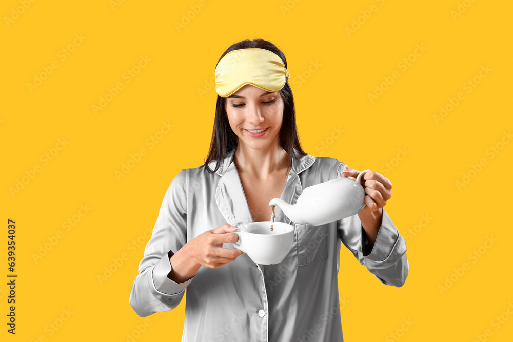 Young woman in pajamas pouring tea into cup on yellow background Young woman in pajamas pouring tea into cup on yellow background