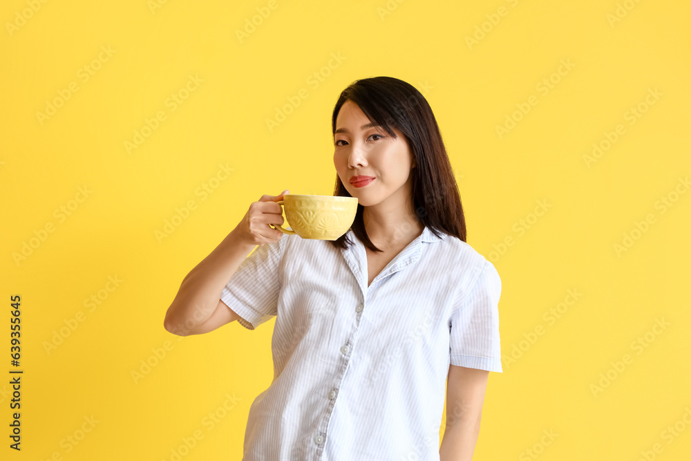 Beautiful Asian woman in pajamas with cup of coffee on yellow background Beautiful Asian woman in pajamas with cup of coffee on yellow background