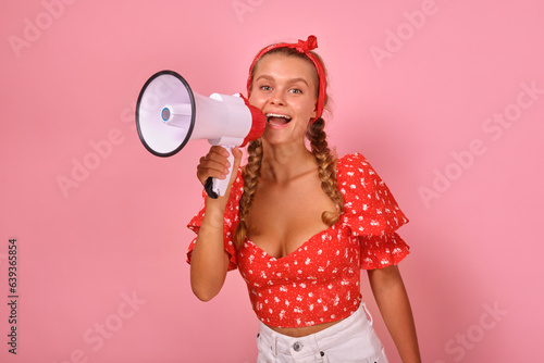 Young cheerful casual Caucasian woman student holds white megaphone in hand and shouts to inform campus residents about approach of holiday party or important exam stands on pink background.