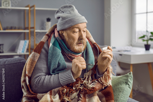 Canvastavla Sick bearded senior man with very high temperature sitting in blanket and hat on couch at home and looking with surprised face expression at thermometer in his hand
