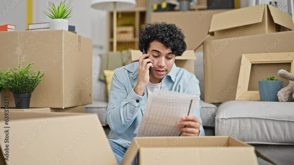 Young latin man reading document talking on smartphone looking upset at new home Young latin man reading document talking on smartphone looking upset at new home