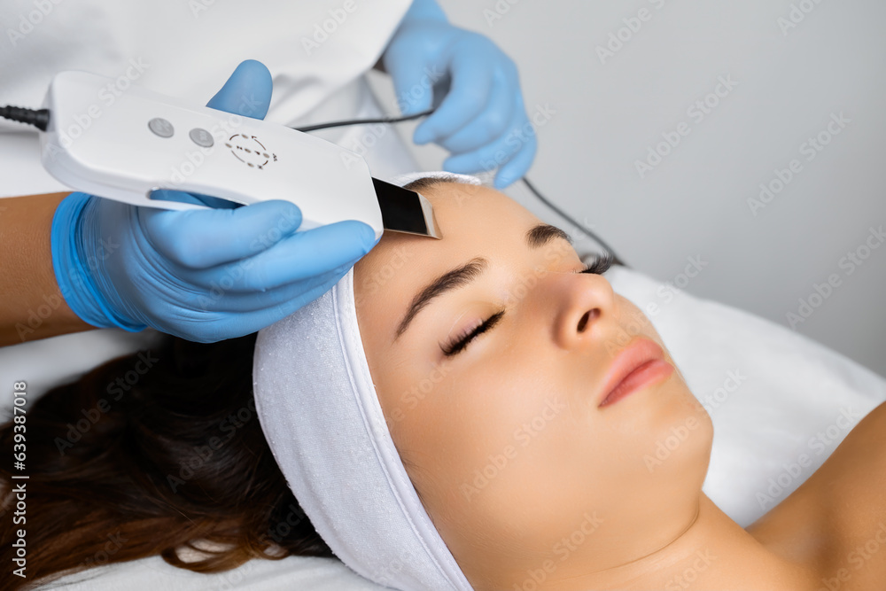 Spa worker in rubber gloves cleans face with ultrasonic device. Specialist does cleansing of body part. Smooth skin effect and blemish removal Spa worker in rubber gloves cleans face with ultrasonic device. Specialist does cleansing of body part. Smooth skin effect and blemish removal