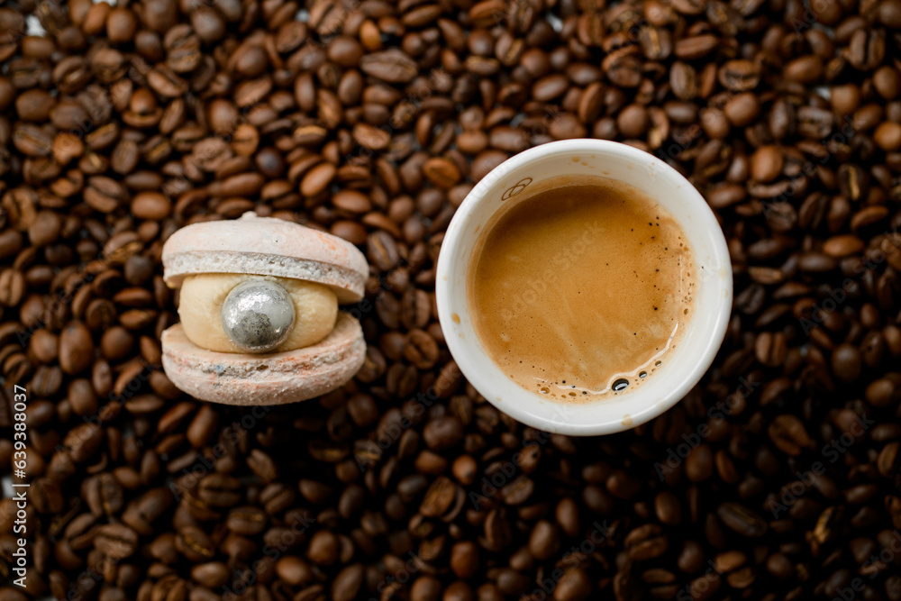 Fototapeta premium Selective focus of white cup with coffee and macaroon on top of fresh roasted coffee beans