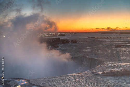 Panoramic view over the Niagara Falls, ON, Canada during winter sunrise with orange sky, water spray from the falls and International Control Dam in distance