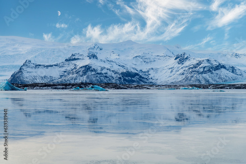 Panoramic view over the mountains surrounding the frozen glacial lagoon of the Jokulsarlon glacier  Iceland with in forefront various blue arctic icebergs and sky with feather clouds and quarter moon