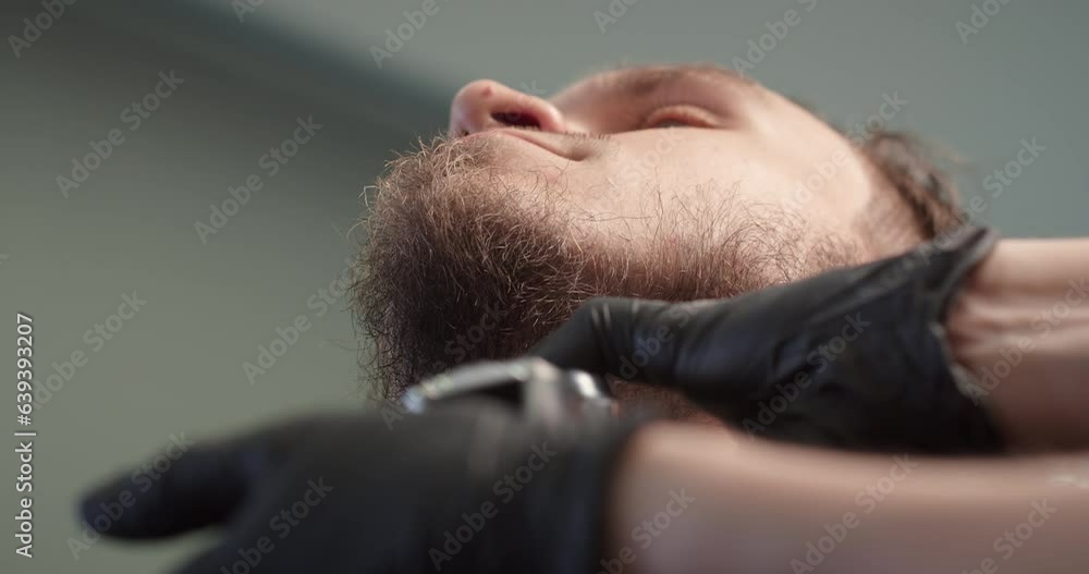 Lower angle shot barber trimming bearded man with shaving machine in barbershop. Hairstyling process. Close-up of a Hairstylist cutting the beard of a bearded male. 4K Lower angle shot barber trimming bearded man with shaving machine in barbershop. Hairstyling process. Close-up of a Hairstylist cutting the beard of a bearded male. 4K