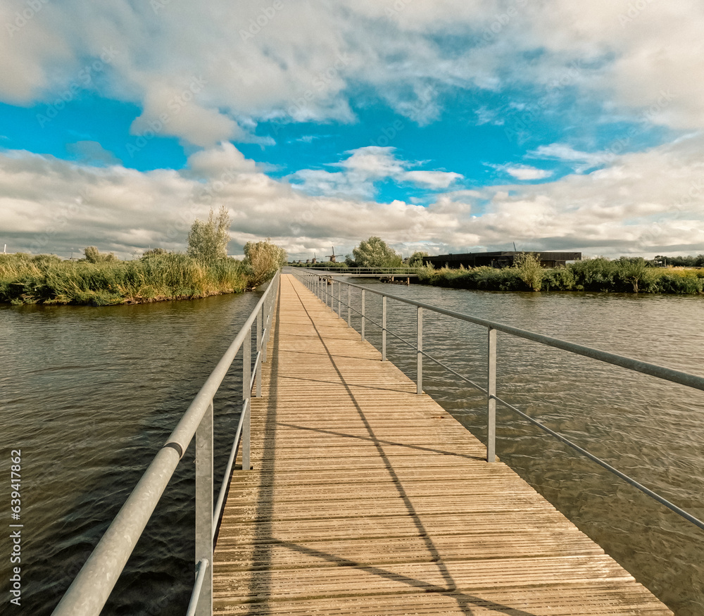 Fototapeta premium Footbridge over the canal system in Kinderdijk, Netherlands. In the distance, the iconic and historical Dutch windmills can be seen. Kinderdijk has been a UNESCO World Heritage Site since 1997.