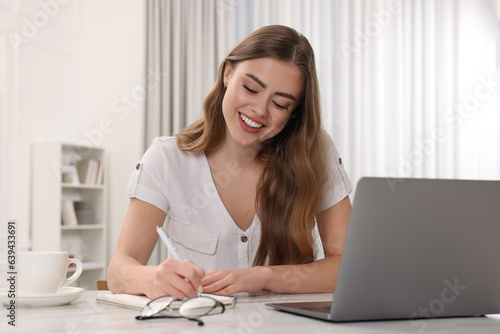 Happy woman writing something in notebook near laptop at white table in room