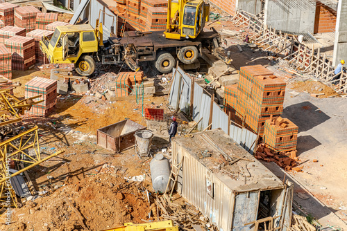 Aerial view of a messy construction site with unrecognizable people picking up red brick blocks using a crane mounted on a truck. an old container, a tank and a crane dismantled on the ground.