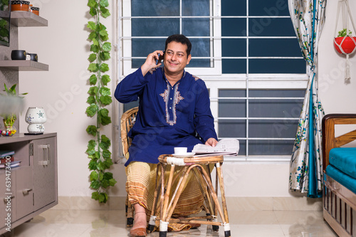 Indian man having tea or coffee and reading news paper while talking to someone on mobile phone at home
