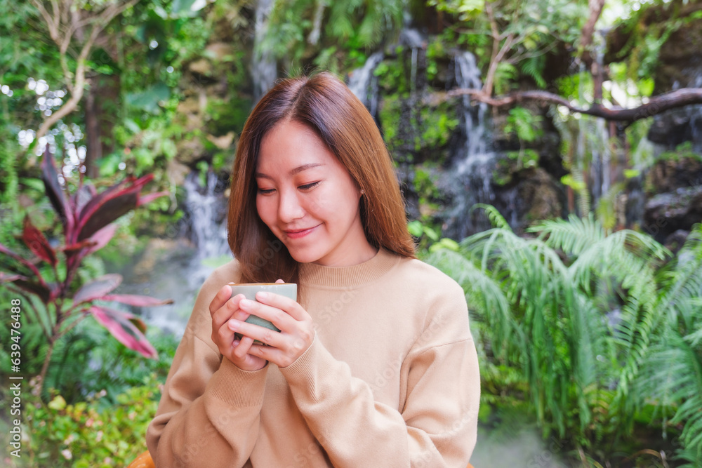 Portrait image of a young woman holding and drinking coffee while sitting in the garden with waterfall