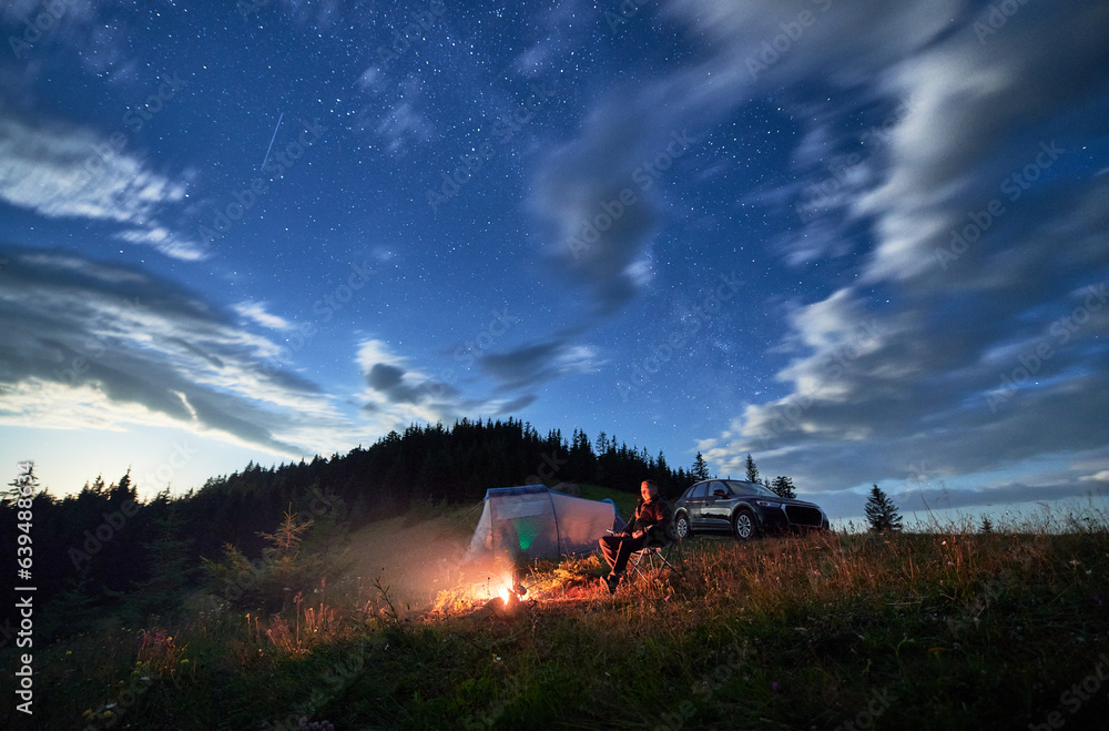 Fototapeta premium Camping in mountains under evening cloudy sky. Young man having a rest next to campfire, near tourist tent and off-road car on hill. Travelling man sitting and stargazing at night.
