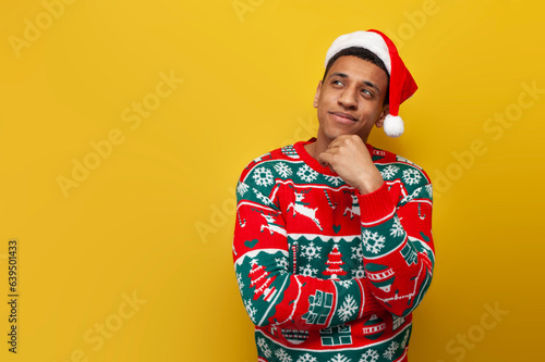 young pensive guy in christmas clothes and santa hat thinks and imagines on yellow isolated background
