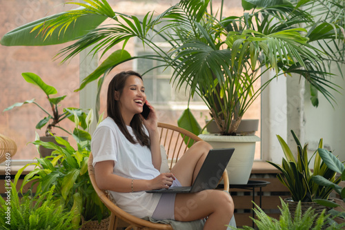 smiling brunette woman talking on the phone and using a laptop while sitting on a comfortable armchair. Remote work concept, lifestyle