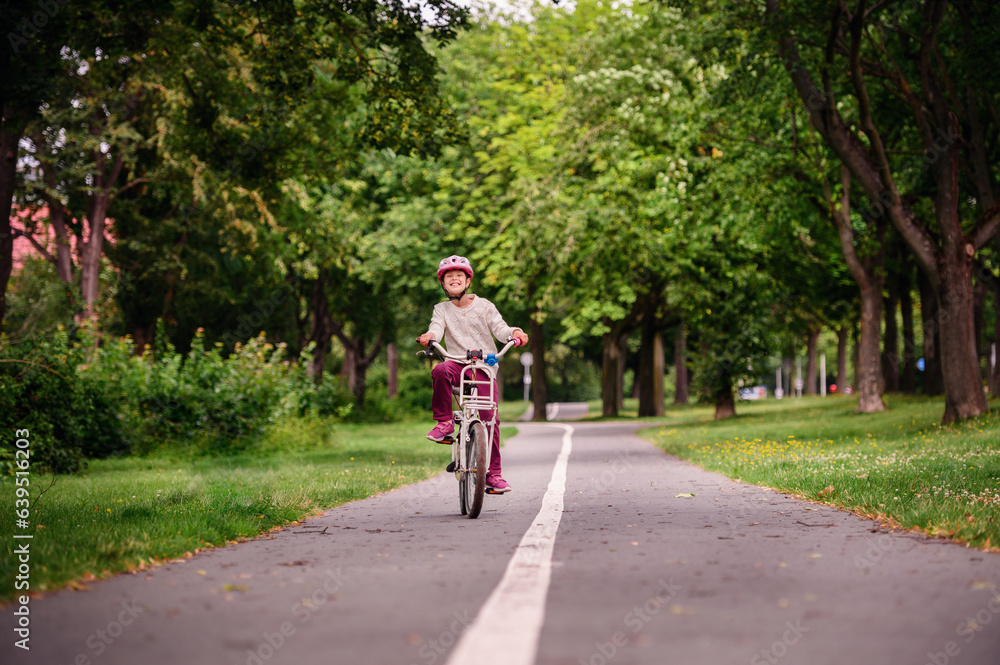 Little schooler girl riding bike in parks. Summer time, wearing helmet. From the back.