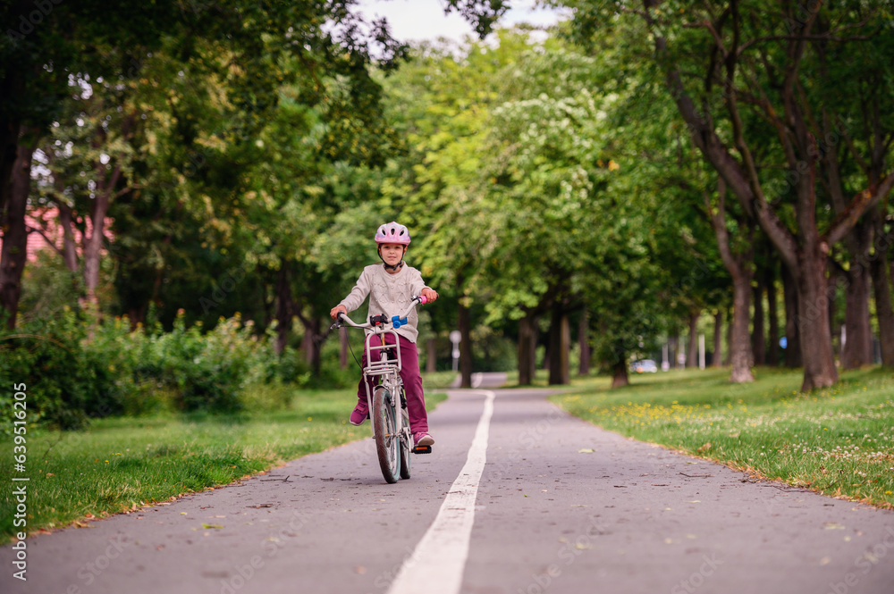 Little schooler girl riding bike in parks. Summer time, wearing helmet. From the back.