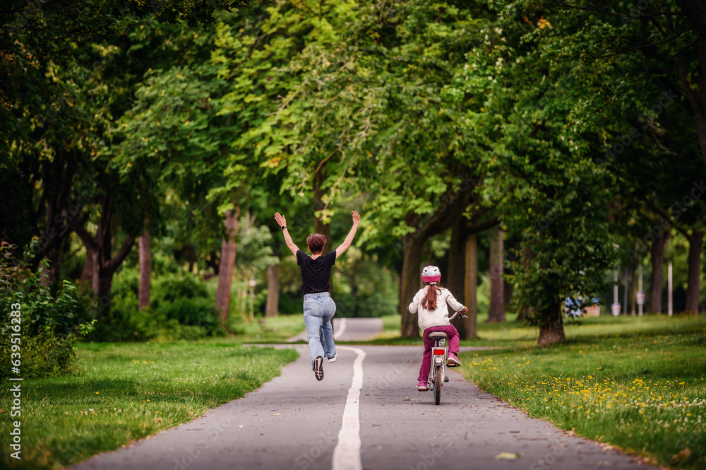 Young mother in jeans and black t-shirt running and playing with her daughter in summer parks while girl riding a bike