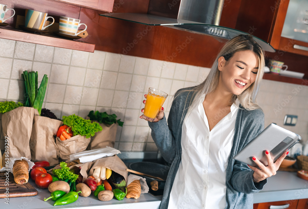 Cheerful young woman using digital tablet while drinking orange juice in the kitchen after returning from grocery shopping.