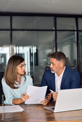 Mature 50s age Latin business man mentoring mid age European business woman discussing project with documents in office. Colleagues, group of partners, professional business people working together.