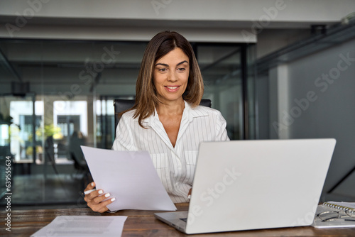 Smiling European Latin business woman accountant analyst holding documents, work at laptop computer doing online trade market tech research. Focused Hispanic businesswoman with paperwork in office. © Stock 4 You