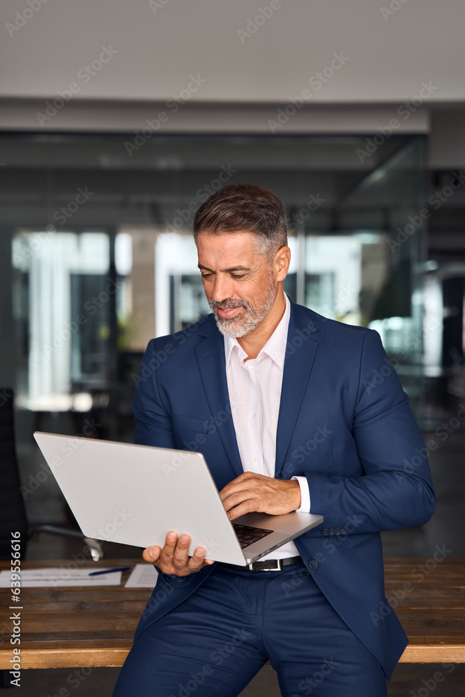 Vertical portrait of mature Latin business man ceo trader using laptop computer,typing, working online in modern office. Middle-age Hispanic handsome businessman entrepreneur looking at laptop at desk