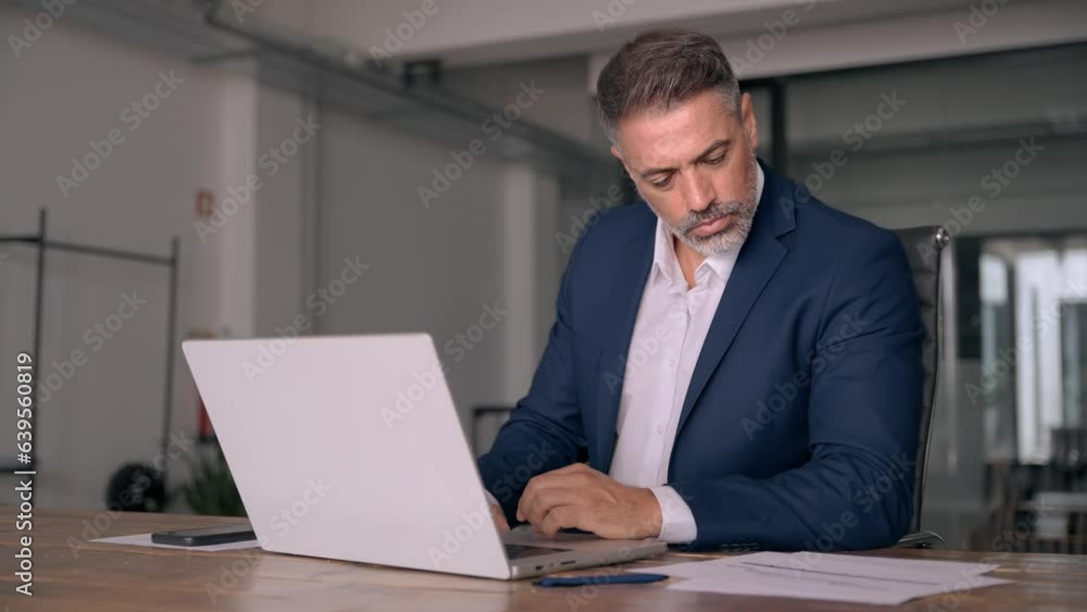 Portrait of mature Indian or Latin business man ceo trader using laptop computer, typing, working in modern office. Middle-age Hispanic smiling handsome businessman entrepreneur looking at laptop. Portrait of mature Indian or Latin business man ceo trader using laptop computer, typing, working in modern office. Middle-age Hispanic smiling handsome businessman entrepreneur looking at laptop.