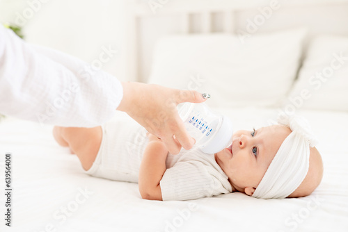 mom's hand feeds the baby from a bottle on the bed in a bright bedroom, the concept of healthy baby food, the baby eats from a bottle