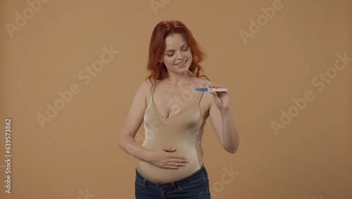 A pregnant woman holds a pregnancy test with a positive result in a studio on an orange background. A happy woman holds her big belly with her hand. photo