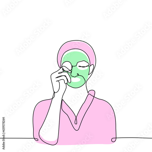 woman in a terry robe and headband removes a green mask from her face with a cotton pad - one line art vector. concept beauty treatments at home, body care