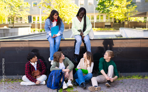 Students are sitting outdoors in the campus. Young people speaking together, reading book and communicating while sitting on the wall.
