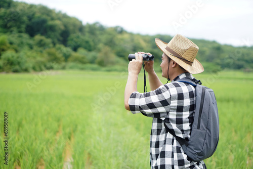 Tela Asian man naturalist wears hat, plaid shirt, backpack, uses binocular to explore nature at paddy field