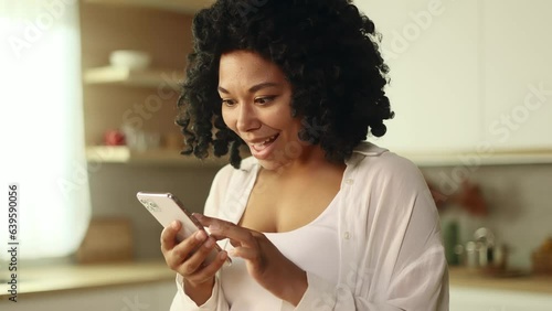 Portrait of young curly woman looking at smartphone cellphone screen with nod of approval and looking at camera pointing at the phone at home Advise Approve Recommend Choice concept